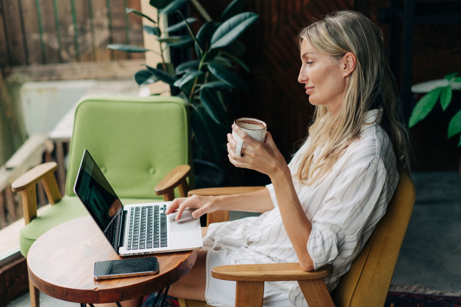 Freelancerin sitzt mit einem Kaffee in der Hand in einem Cafe und arbeitet am Laptop.