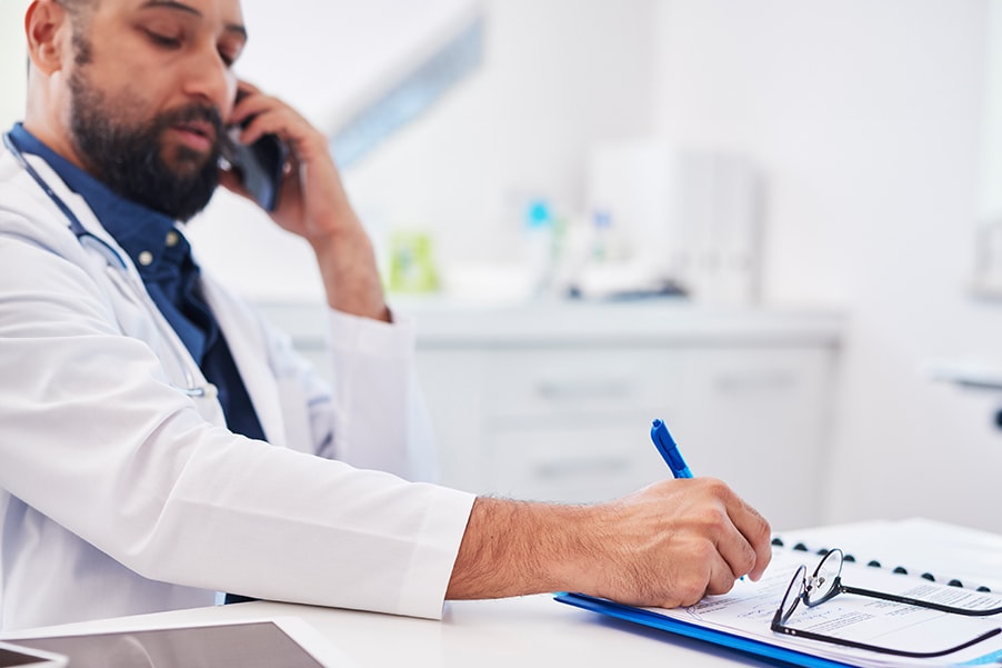 Telefonische Krankmeldung durch einen Arzt. Er telefoniert im Arztzimmer und macht sich dabei Notizen an einem Schreibtisch.