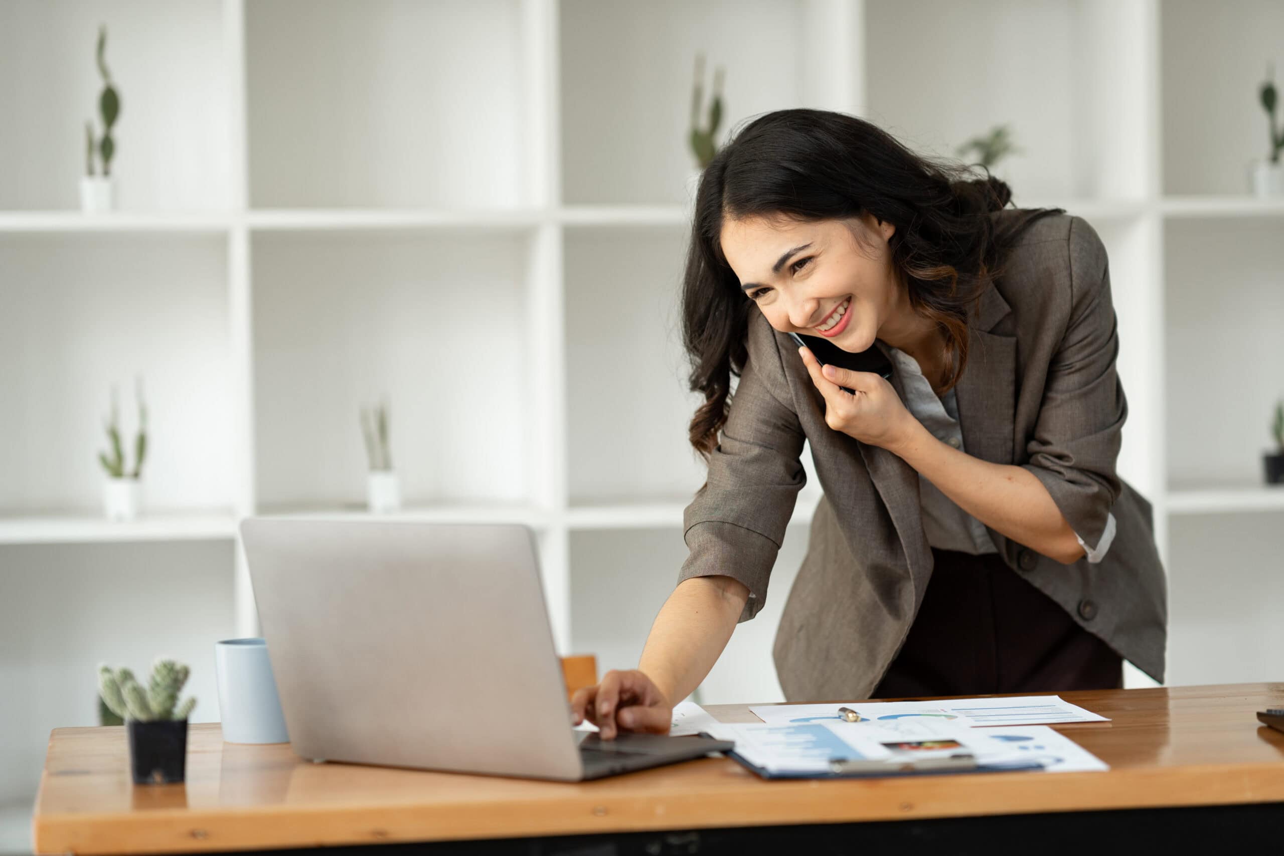 Arbeitnehmerzufriedenheit dargestellt durch eine junge glückliche Mitarbeiterin, die telefoniert und parallel am Laptop im Home-Office arbeitet.