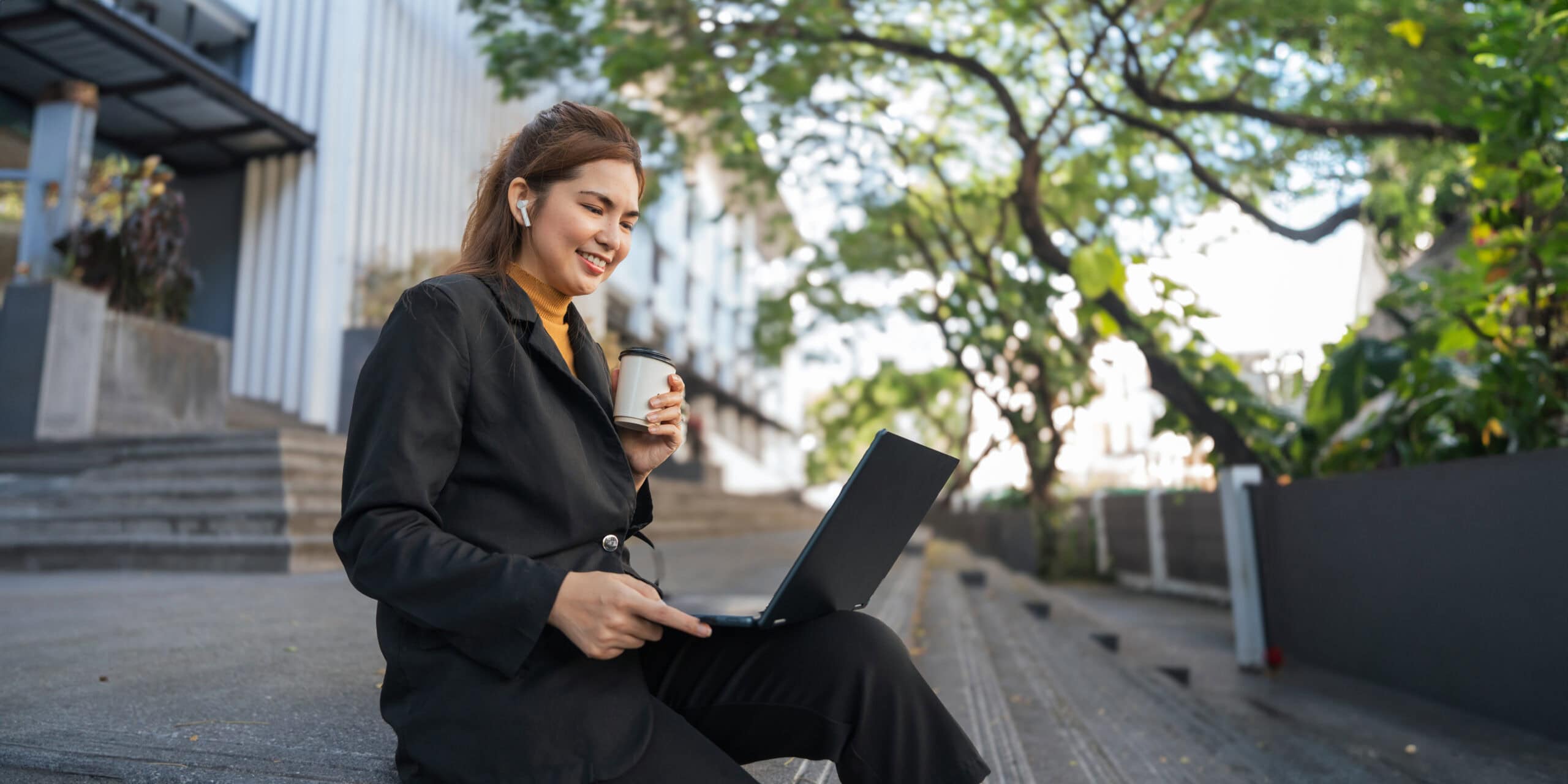 Lächelnde junge asiatische Business-Frau mit Laptop sitzt im Freien beim hybride arbeiten.