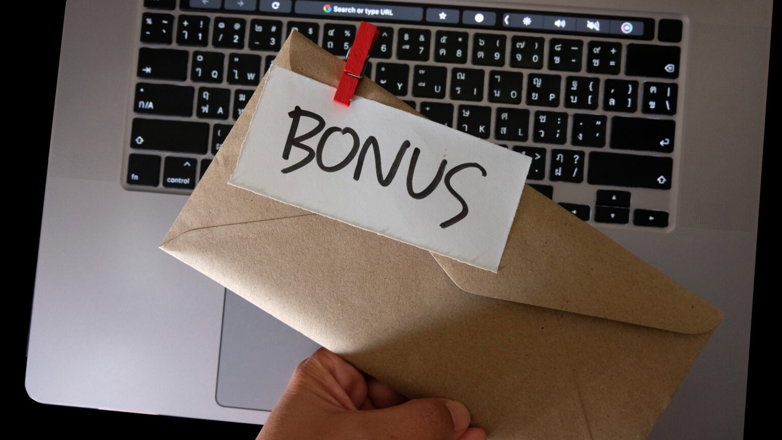 Laptop steht auf dem Tisch. Eine Person hält einen Umschlag in der Hand, an dem eine Notiz mit Bonus draufsteht.