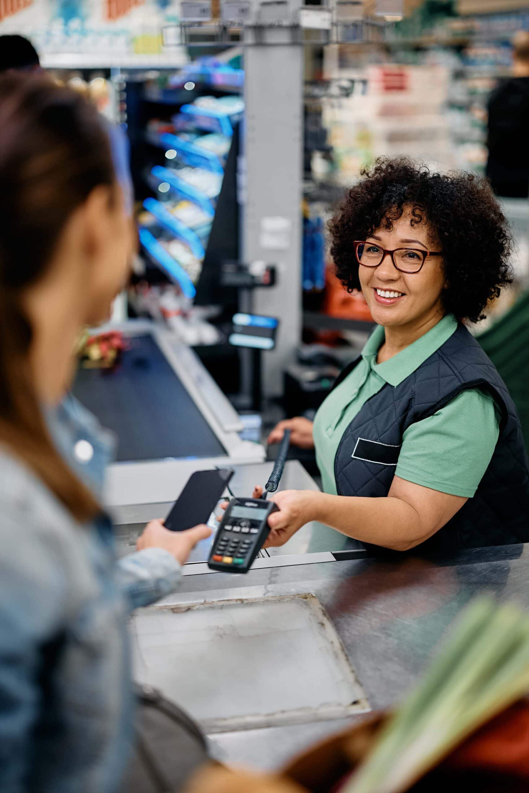 Glückliche Kassiererin hält Kreditkartenleser in der Hand, während der Kunde im Supermarkt mit dem Smartphone bezahlt.