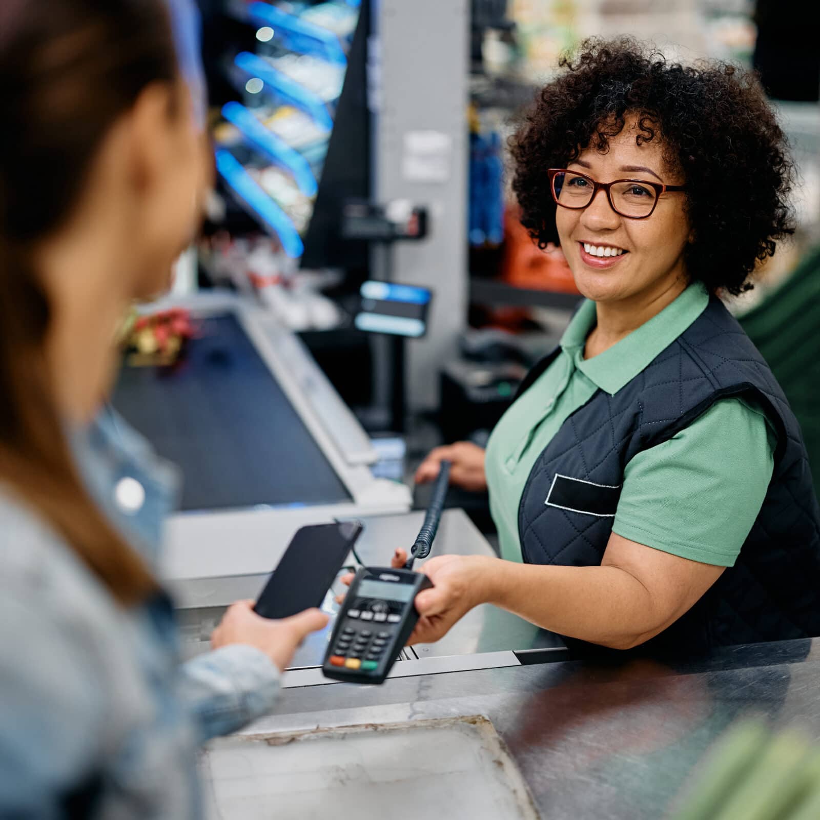 Glückliche Kassiererin hält Kreditkartenleser in der Hand, während der Kunde im Supermarkt mit dem Smartphone bezahlt.