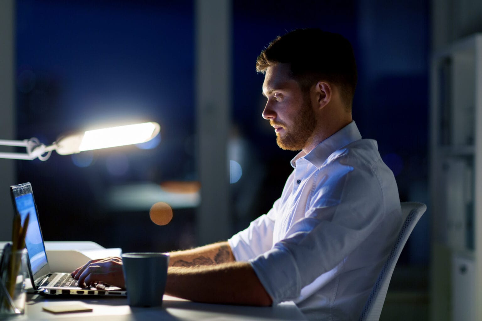 Mann sitzt mit einer Tasse Kaffee am Schreibtisch und arbeitet nachts im Büro am Laptop